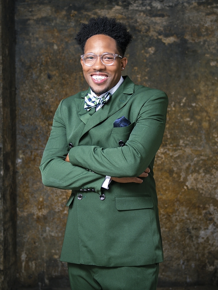 Mitch Mitchell, a young Black man wearing a green suit with a bow tie and glasses, smiles for a head shot, crossing his arms.